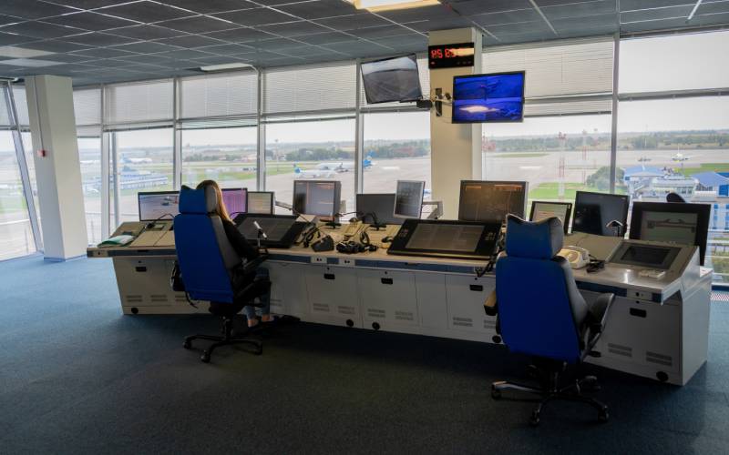 Two flight dispatchers sit in heavy-duty ergonomic chairs in an airport control center. A window shows planes on a runway.