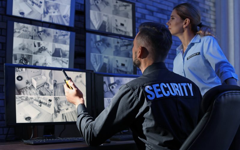 Two security guards wearing security gear looking at multiple monitors on top of a desk and mounted on a wall inside a room.