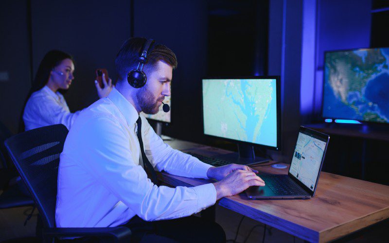 Man wearing a white shirt and a headset sitting on a chair using a computer inside a dispatch center with coworker beside him.