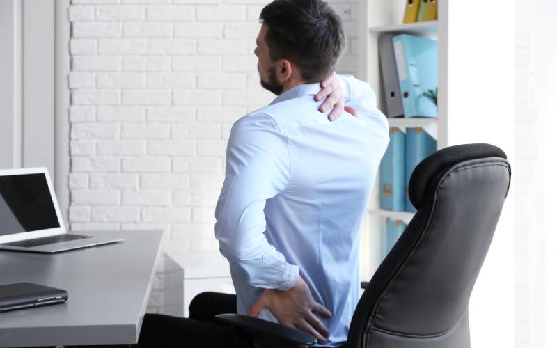 Young man wearing a blue shirt sitting behind an office desk with a laptop stretching and grabbing his neck and lower back.