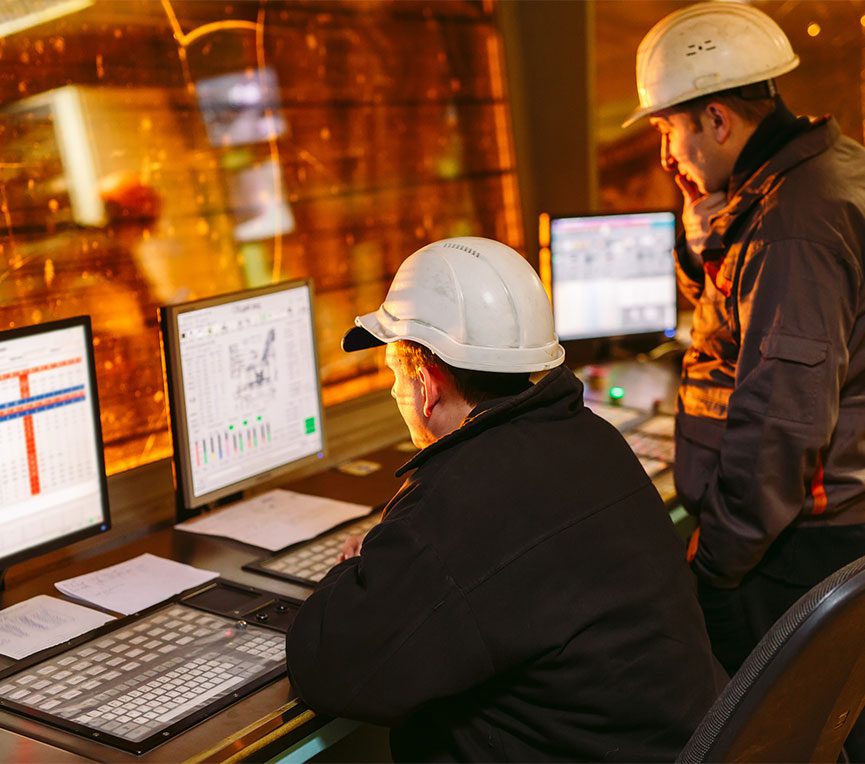 2 men wearing hard hats look at computer screens behind a glass window to a operations floor