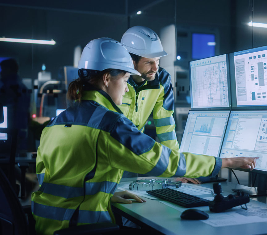 two people wearing hard hats sit at a computer with 6 monitors and look at machine plans