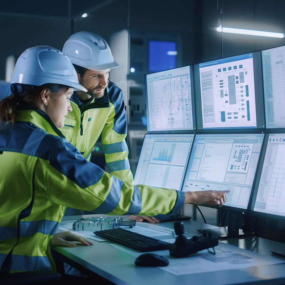 two people wearing hard hats sit at a computer with 6 monitors and look at machine plans